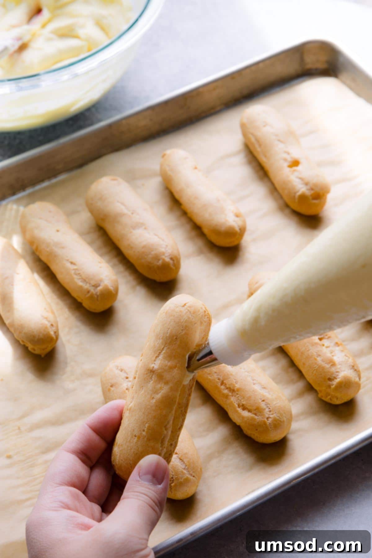 Decadent Chocolate Eclairs 10 A hand demonstrating how to fill an eclair shell by inserting a piping bag into a slit on the side of the pastry.