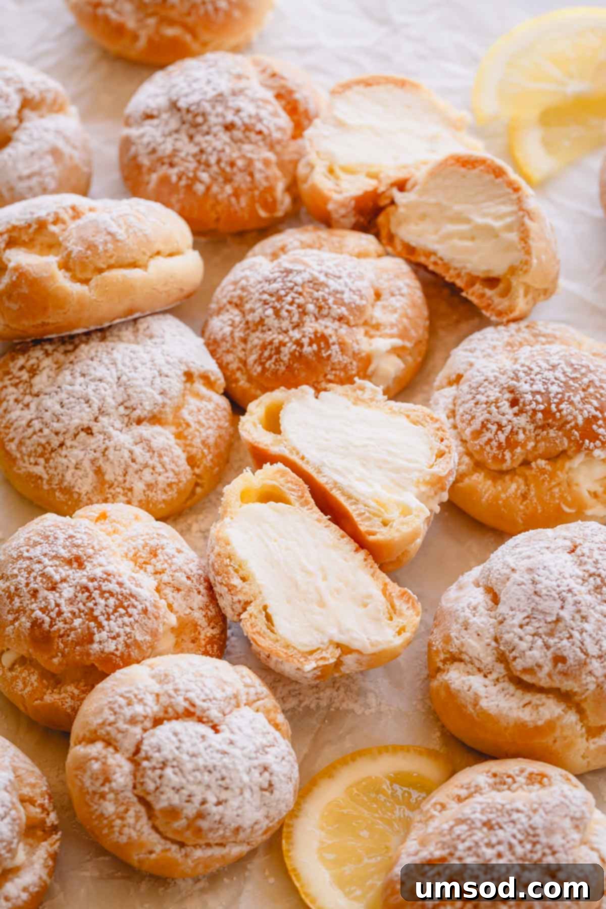 Zesty Lemon Cloud Puffs 16 Overhead image of beautifully presented lemon creme puffs, some dusted with powdered sugar, one split in half revealing its filling.