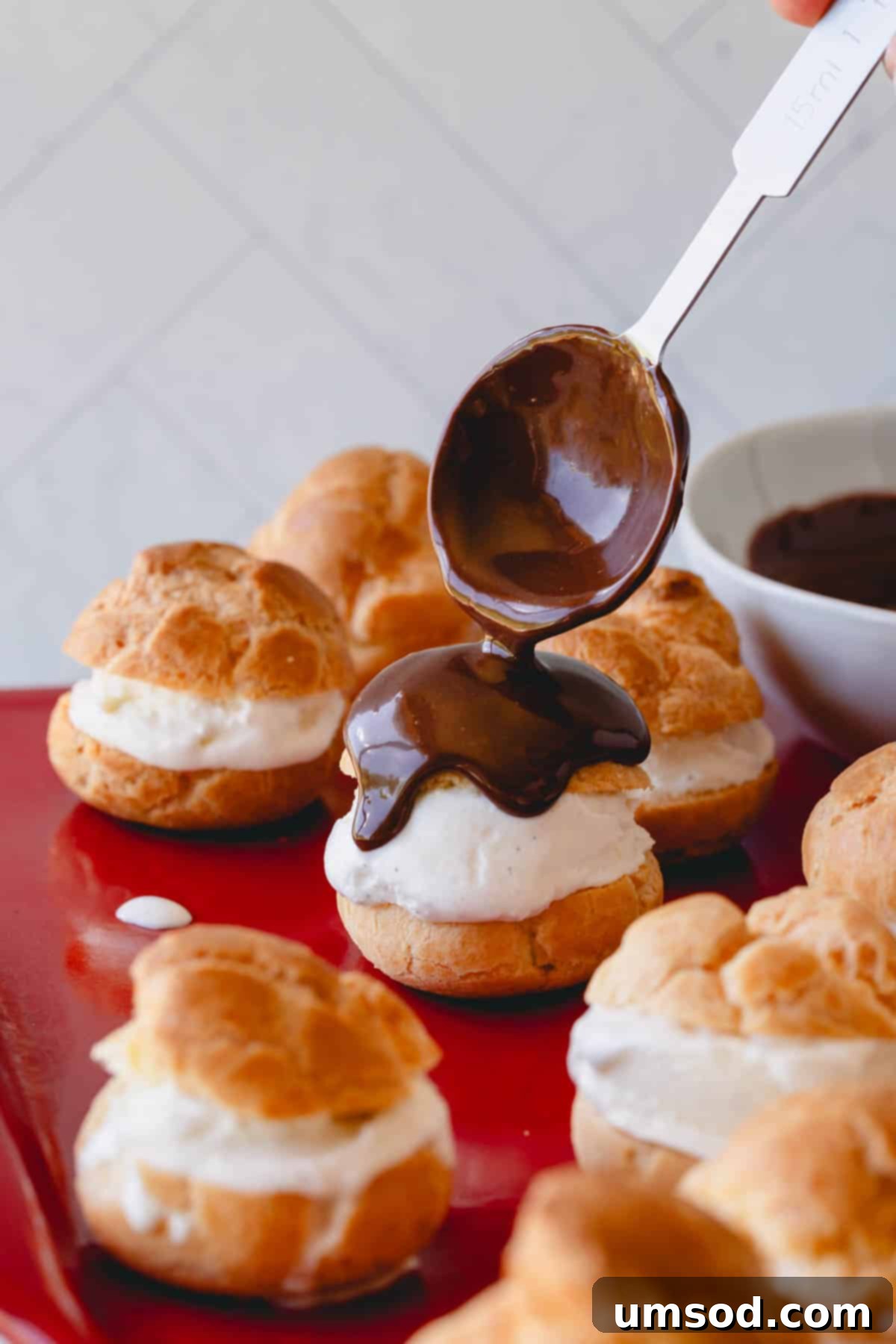 Warm chocolate ganache being gracefully drizzled over a tray of ice cream-filled profiteroles.