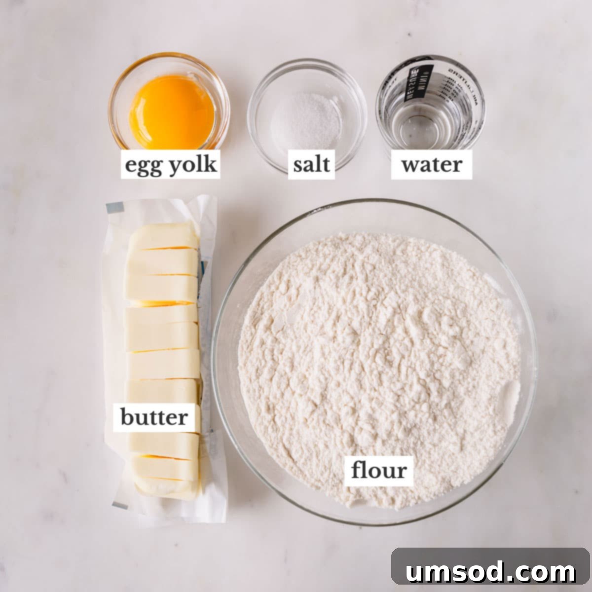 Individual bowls displaying all the necessary ingredients for butter pie crust: flour, salt, cubed frozen butter, egg yolk, and ice water.