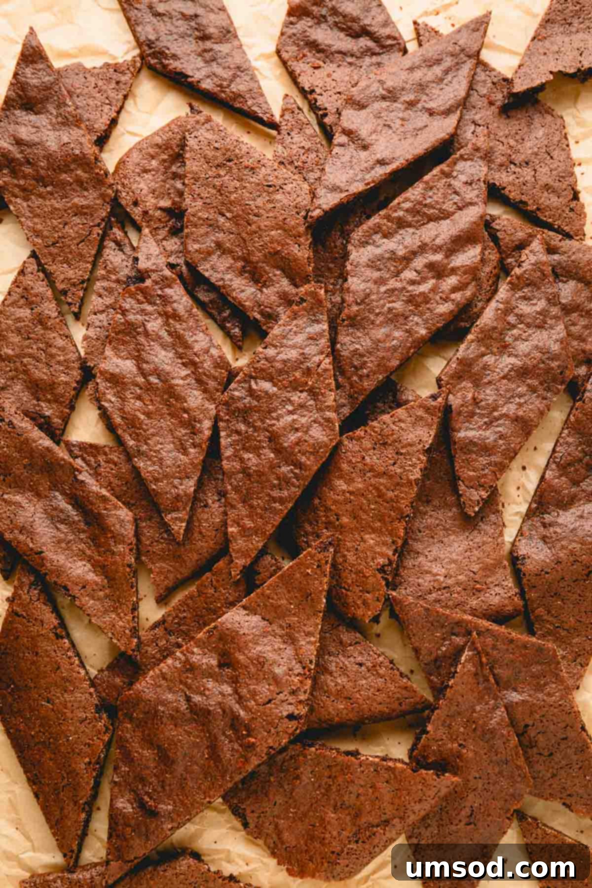 Numerous pieces of broken brownie crisp scattered on a parchment paper-lined baking sheet, ready for serving.