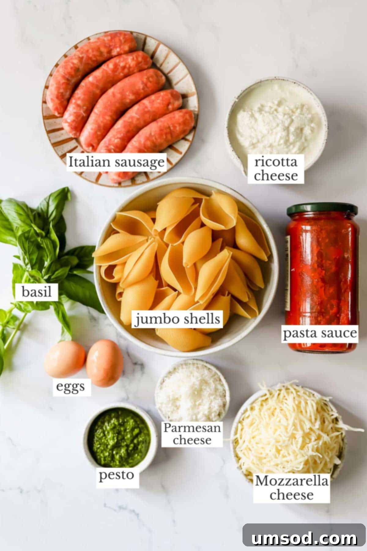 A selection of fresh ingredients including pasta shells, Italian sausage, ricotta cheese, pesto, mozzarella, and eggs, arranged on a rustic wooden board.