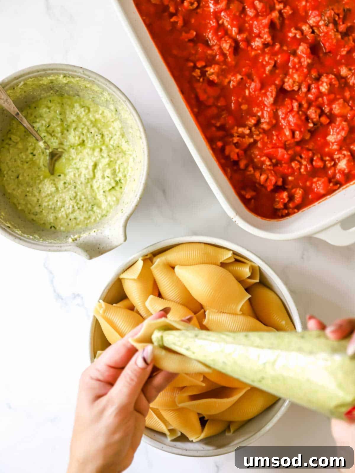 A culinary scene showing a baking dish with marinara sauce, a bowl of pesto, and par-cooked pasta shells, with one shell being expertly stuffed with the pesto ricotta filling.