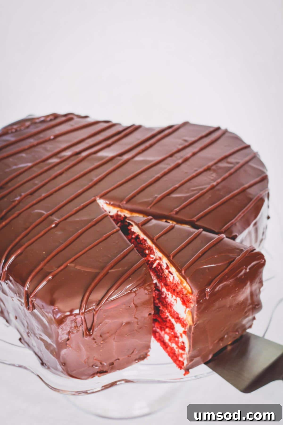 Heart shaped chocolate covered red velvet cake on a glass platter with a slice cut.