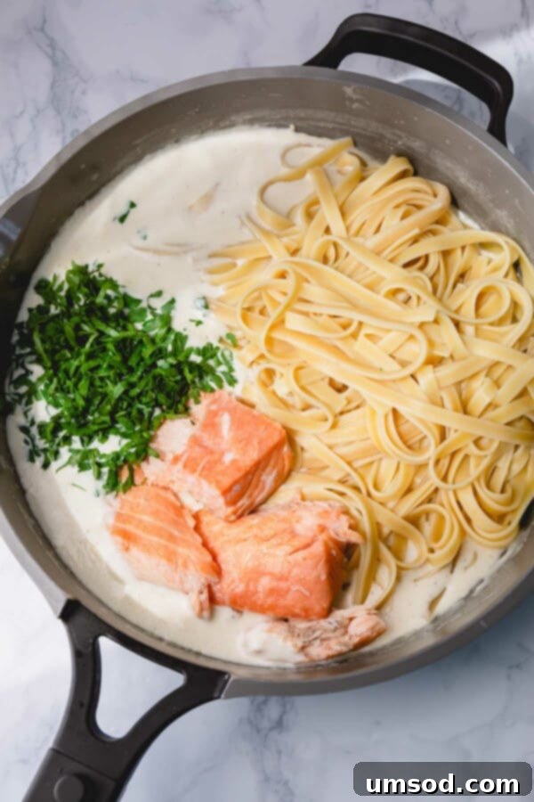 A skillet filled with cooked fettuccine, chunks of hot smoked salmon, and fresh parsley, all tossed in a creamy Alfredo-style sauce.