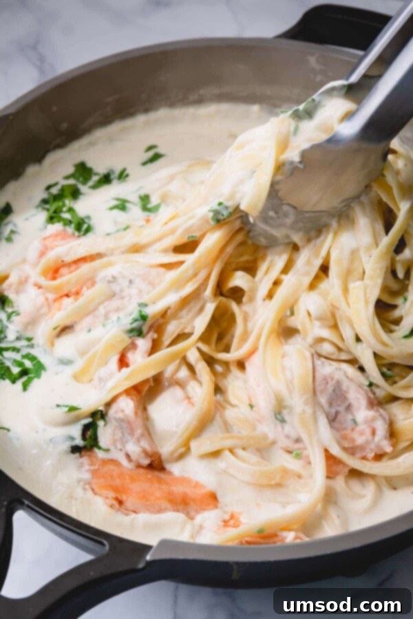 Kitchen tongs expertly tossing fettuccine pasta into the creamy Alfredo sauce in a large skillet, ensuring every strand is coated.