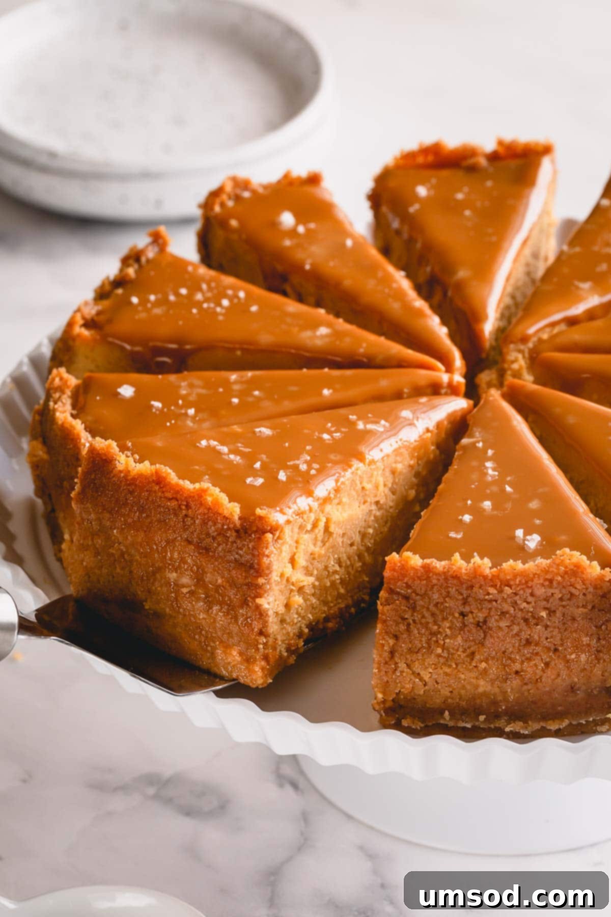 A beautifully sliced Dulce de Leche Cheesecake on a pristine white serving platter, revealing its smooth, creamy layers, rich caramel topping, and crumbly crust.