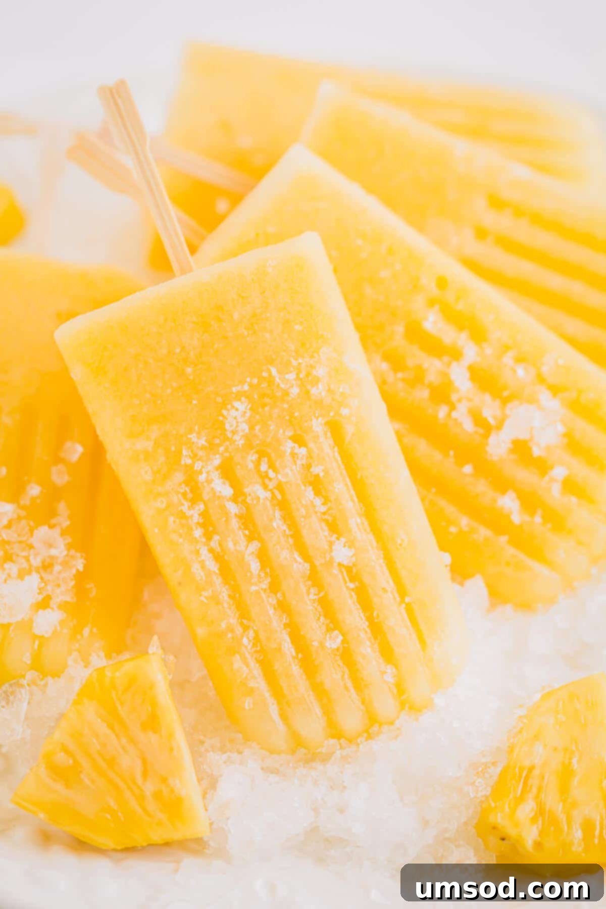 Handful of boozy pineapple popsicles on crushed ice, garnished with mint and lime slices, set against a bright, summery background.