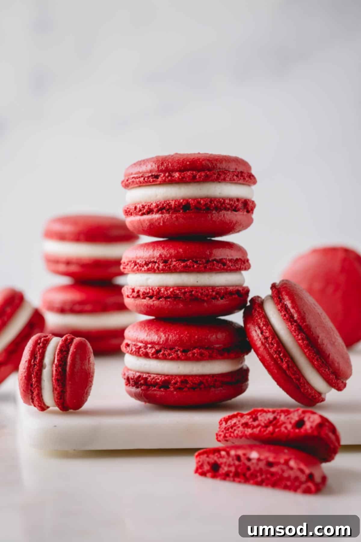 Three perfectly stacked red velvet macarons, brimming with creamy white frosting, surrounded by more macarons on a pristine white surface.