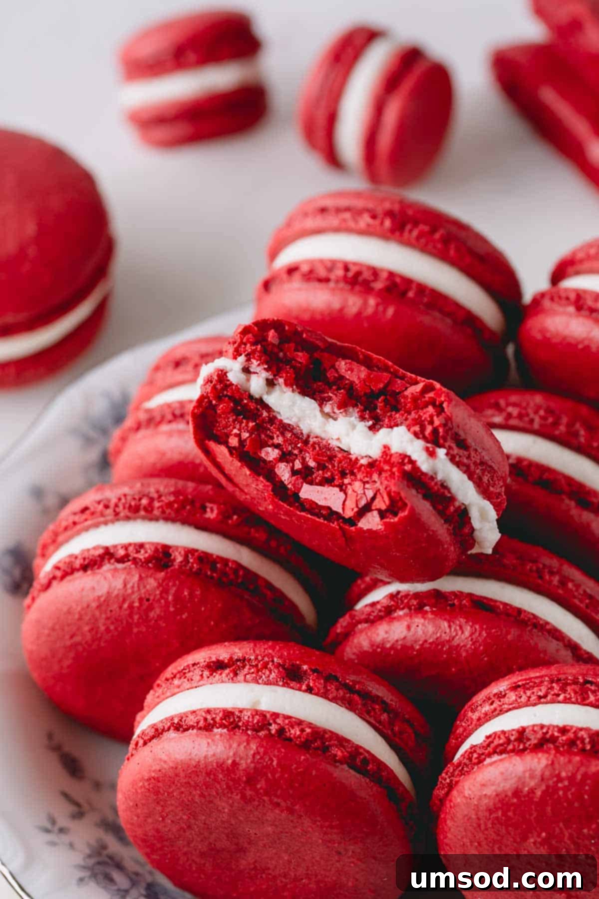 A red velvet macaron on a white plate, with a bite taken out, revealing its rich red shell and white cream cheese filling, surrounded by other perfect macarons.