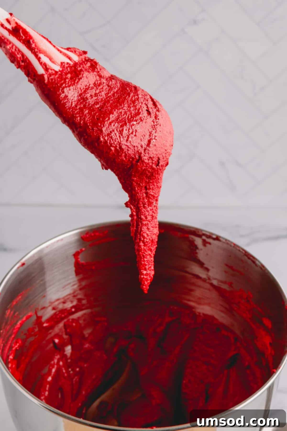 A smooth, red macaron batter gracefully flowing off a spatula, creating a perfect ribbon into the mixing bowl, illustrating the ideal consistency for piping.