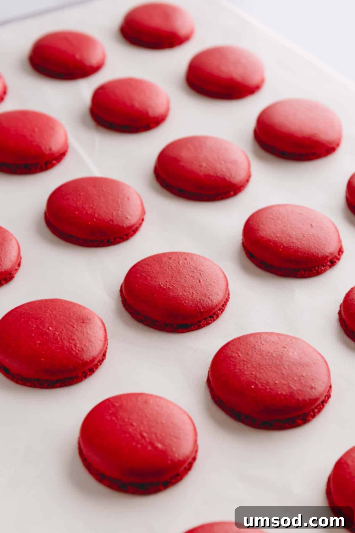 A close-up view of perfectly baked red velvet macaron shells resting on a white Teflon mat, showcasing their smooth tops and delicate 'feet'.