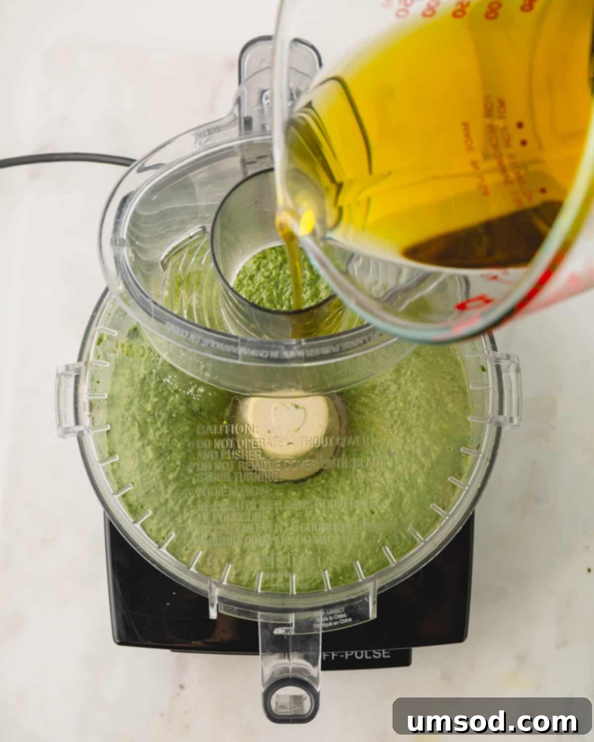 Big Batch Basil Pesto Recipe 11 Olive oil being slowly drizzled into a running food processor containing basil pesto ingredients.